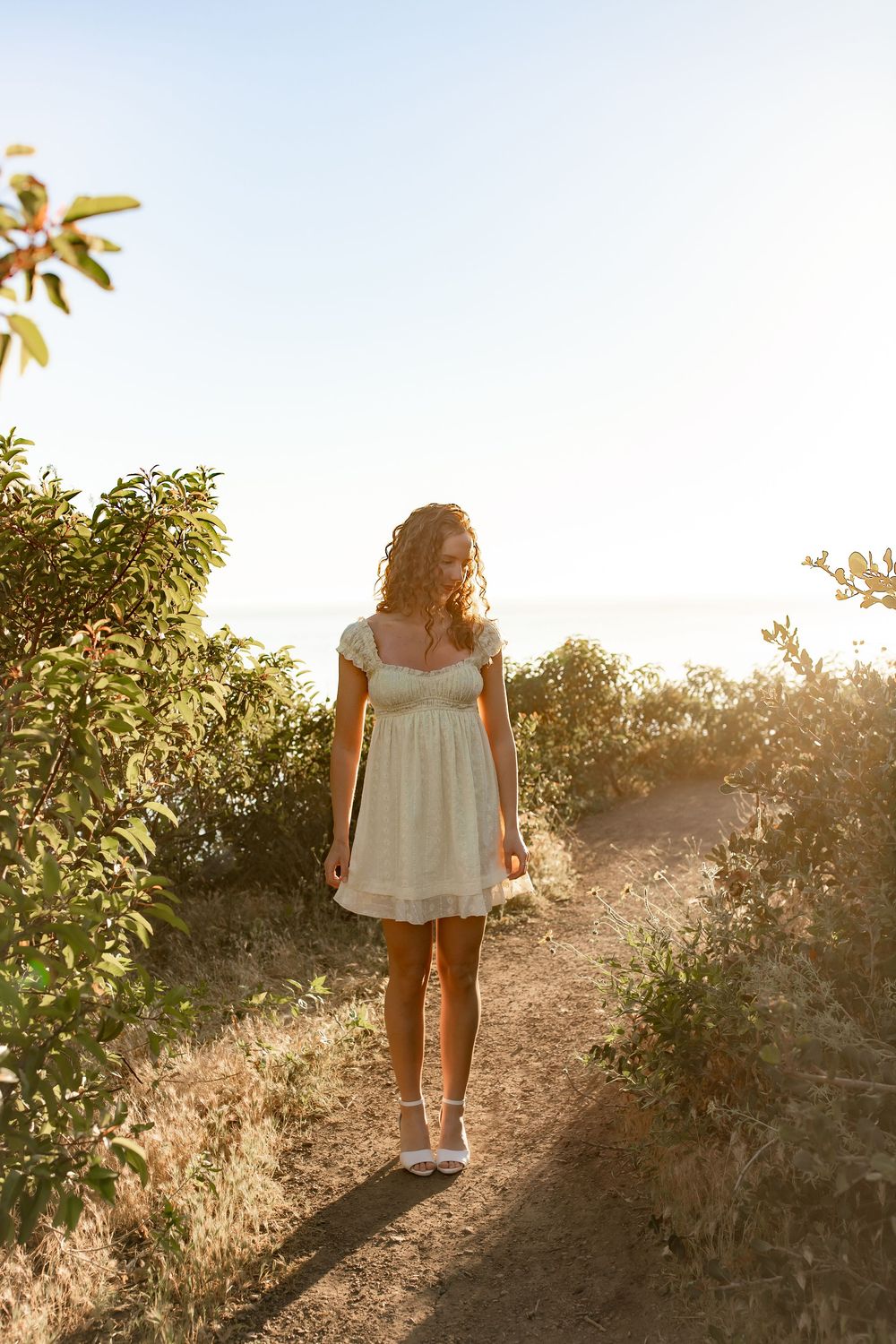 Woman in white sundress walking on a sunlit coastal trail surrounded by green shrubs at golden hour.
