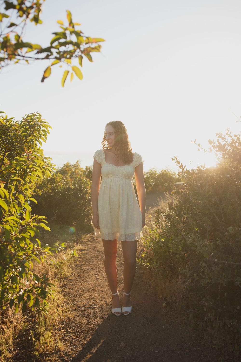 Woman in white sundress walking sun-drenched garden path surrounded by lush green foliage.