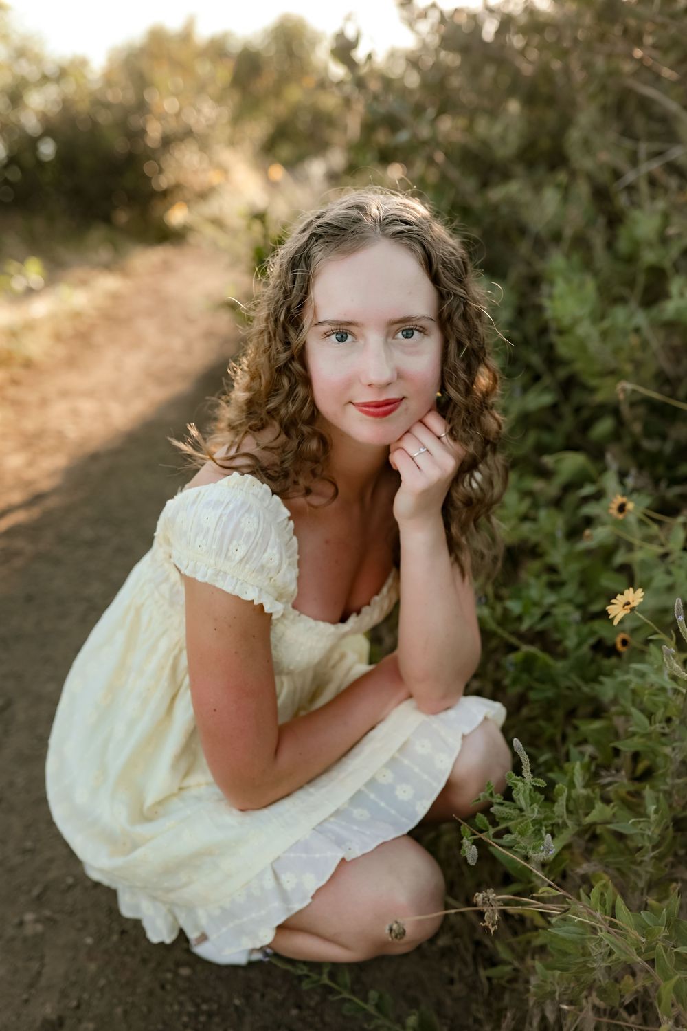 Young woman in yellow dress crouching on a dirt path surrounded by wildflowers and greenery in golden hour light.