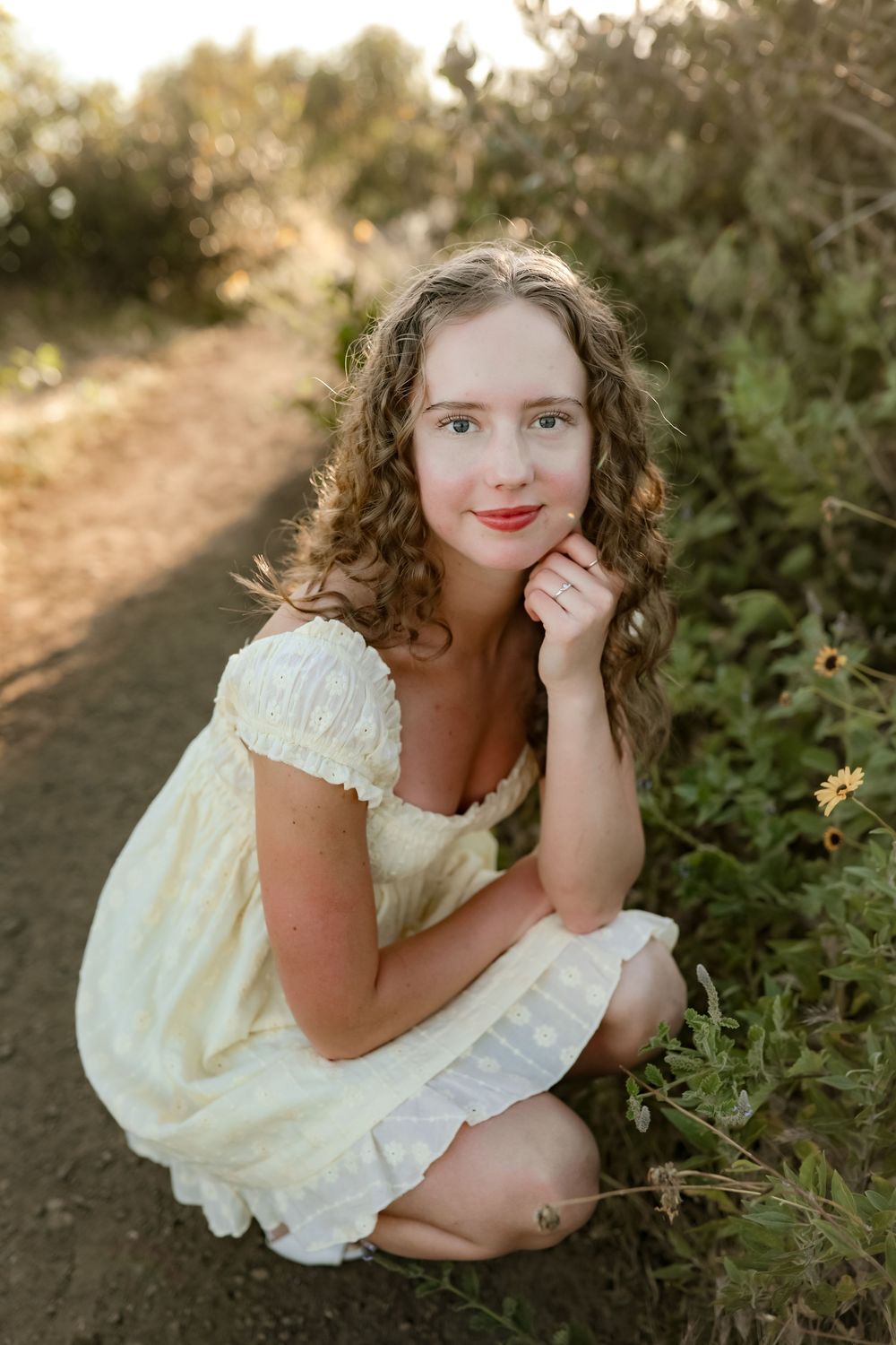 Young woman with curly hair in yellow dress crouching by wildflowers on a sunlit trail.