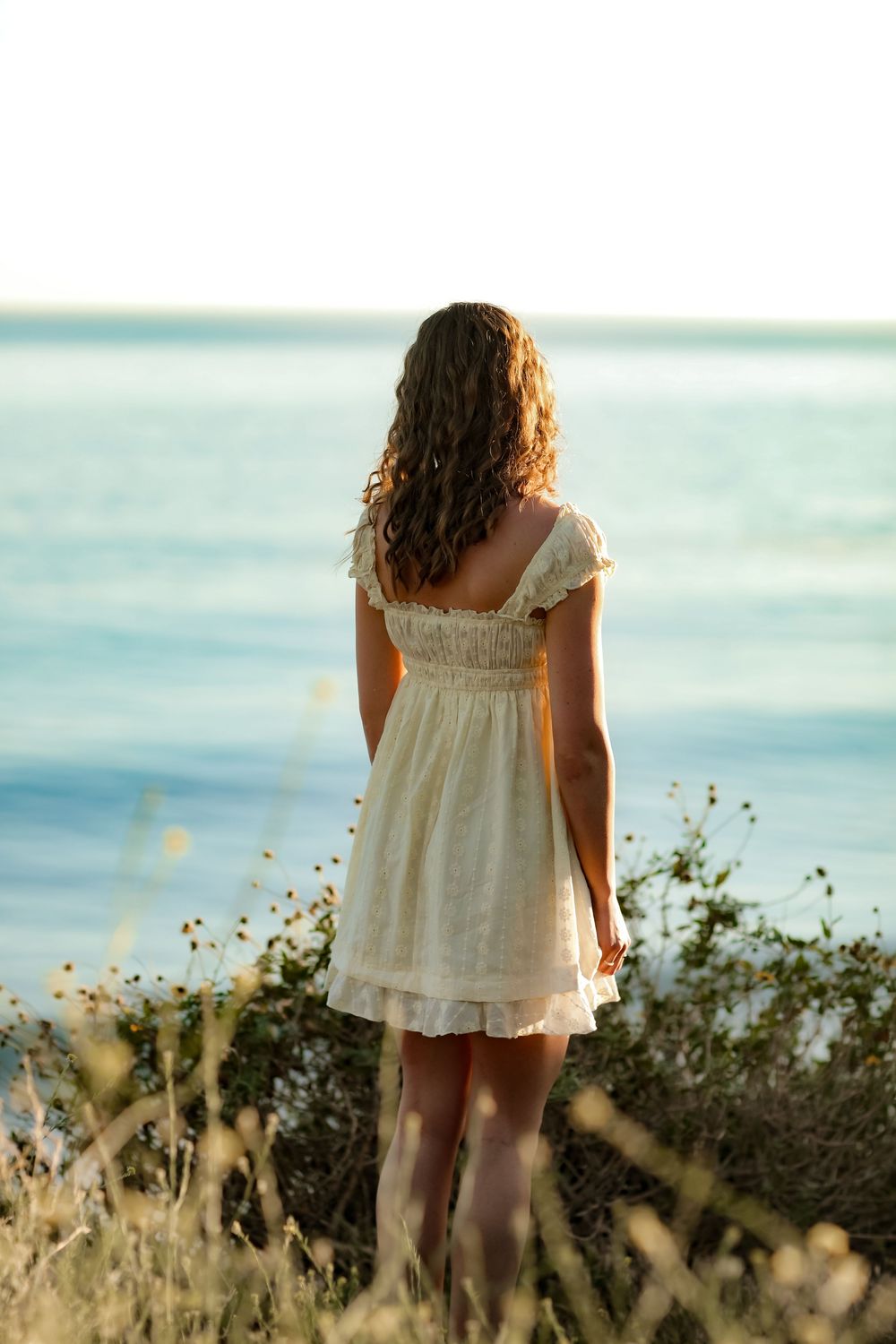 Young woman in white dress standing on coastal cliff overlooking calm blue ocean at golden hour.