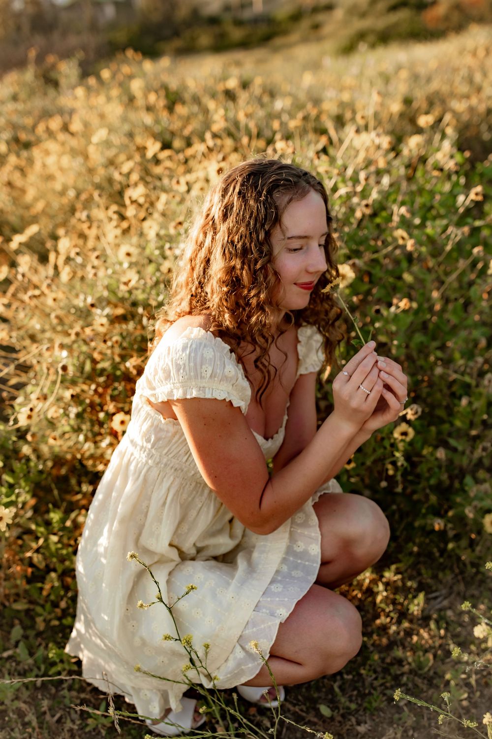 Young girl in white dress crouching in golden field, smelling a small white wildflower at sunset.