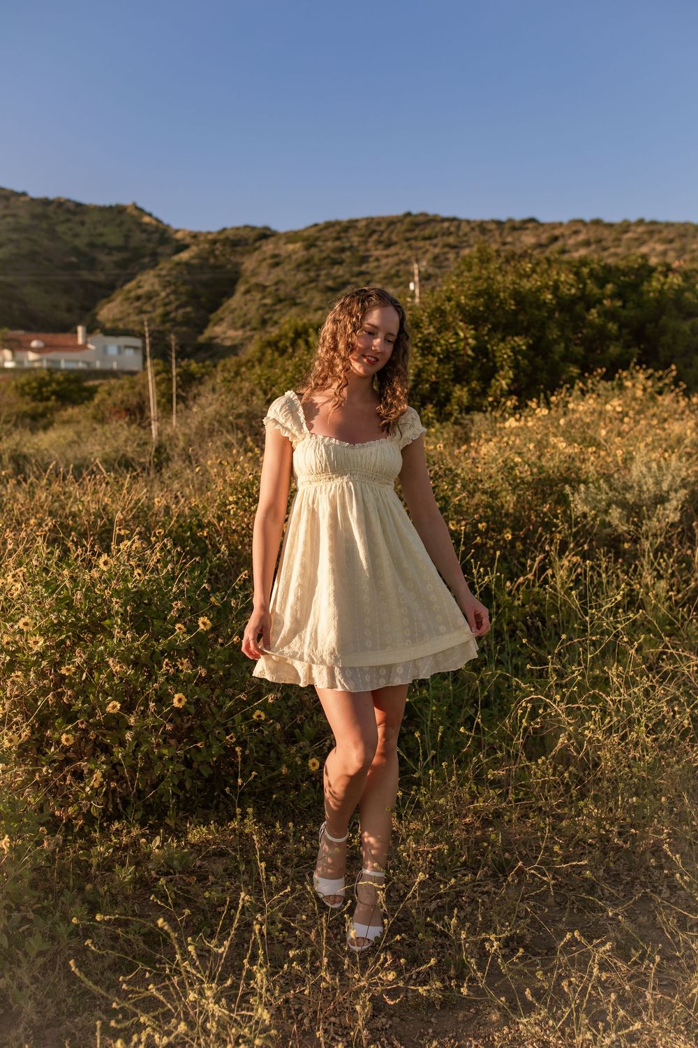 Young woman in cream babydoll dress smiling in golden hour sunlight among wildflowers with green hills behind.