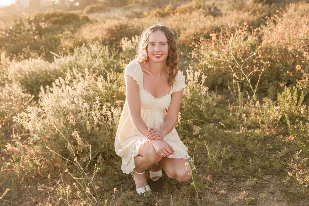 Young blonde woman in yellow dress crouching in golden sunlit field, smiling outdoors portrait photography.
