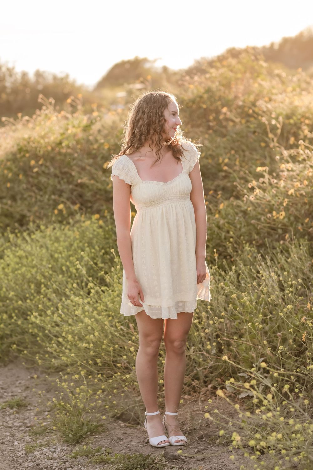 Young woman in cream lace dress standing on a sunlit dirt path surrounded by golden wildflowers.