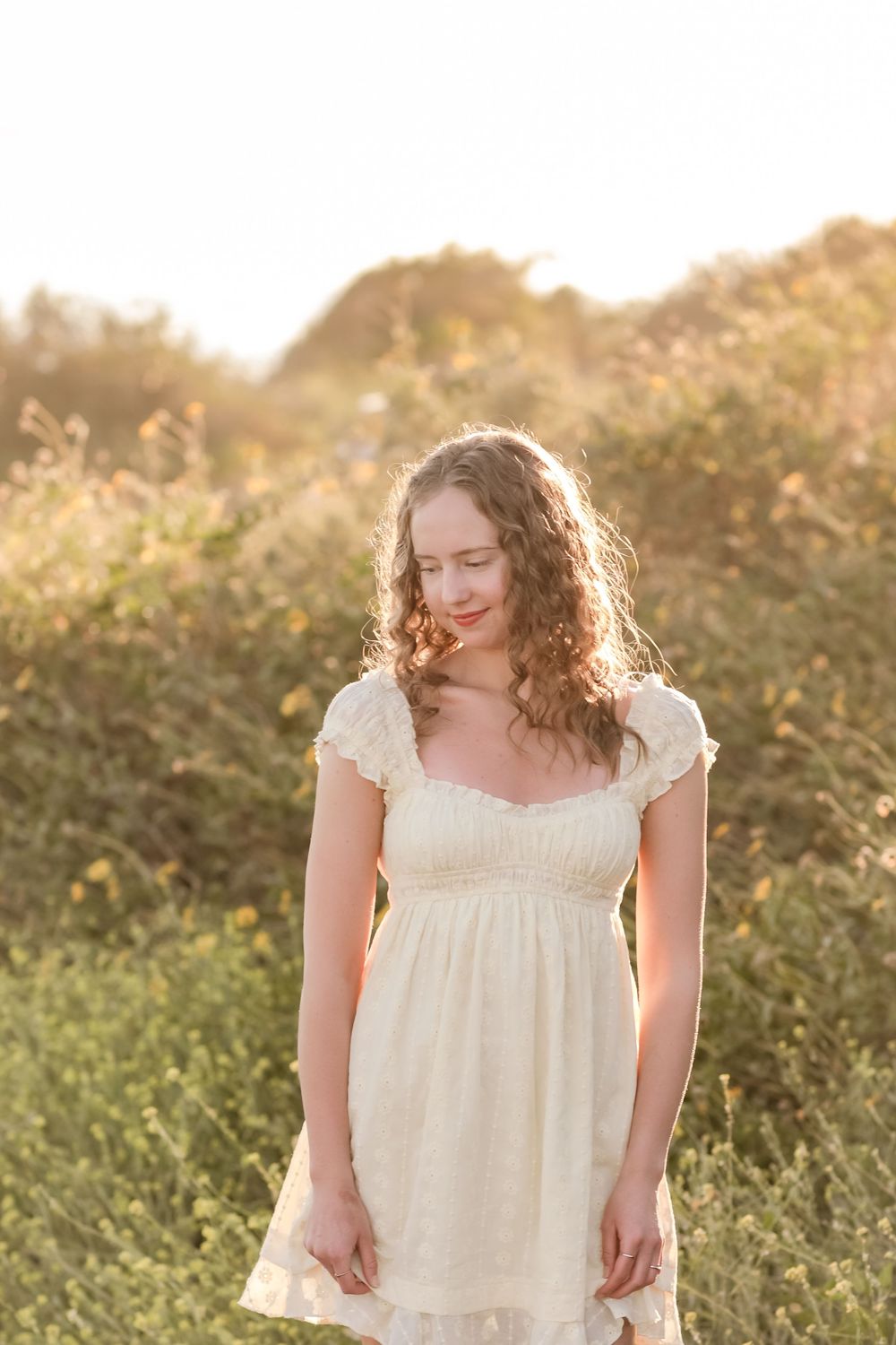 Young woman in white lace dress standing in sunlit field with golden hour light and wildflowers.
