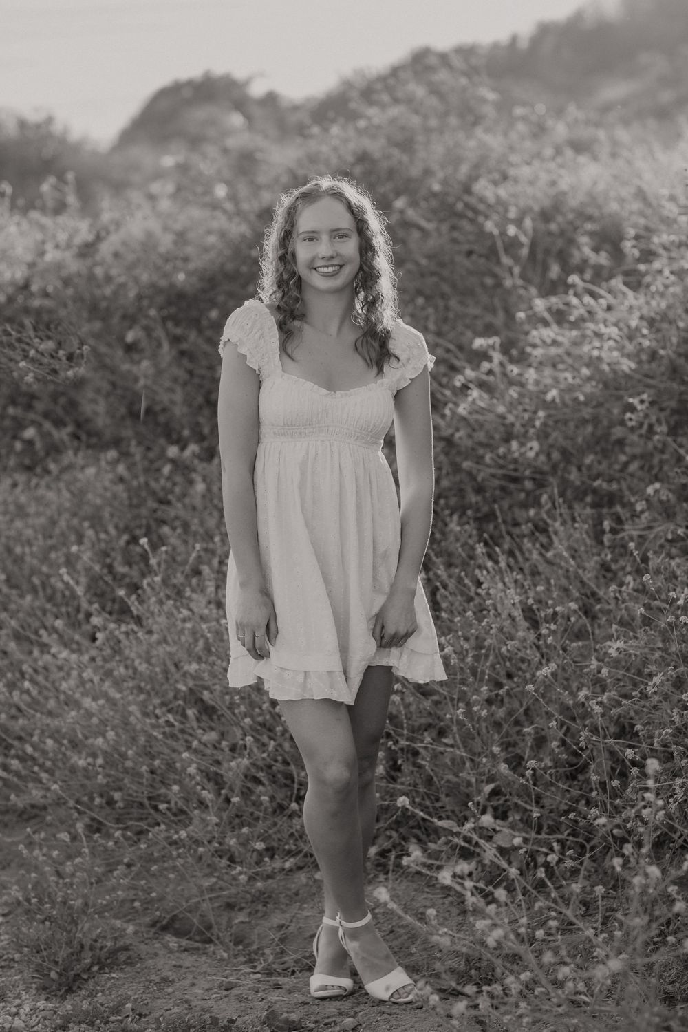 Smiling young woman in white sundress posing in black and white photo among wildflowers.