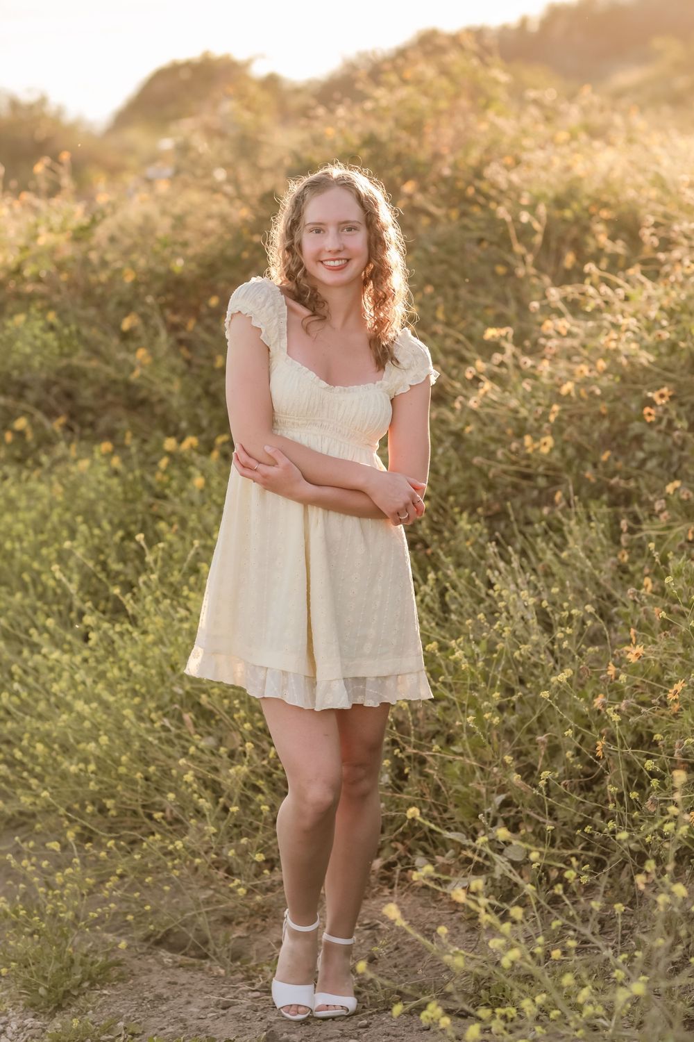 Smiling teen girl in cream lace dress posing among golden wildflowers during sunset.