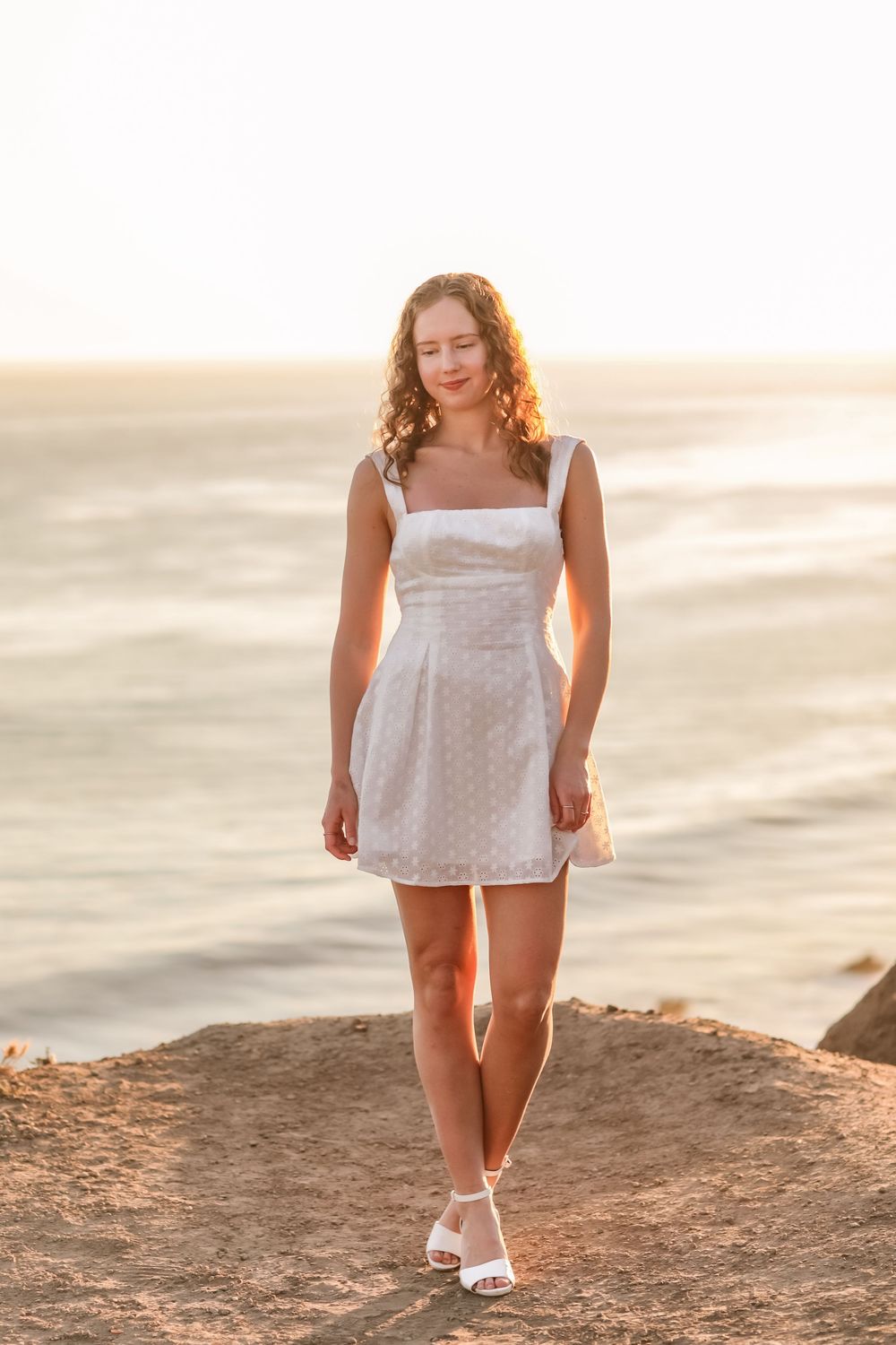 Young woman in white polka dot sundress standing on coastal cliffs with ocean sunset backdrop.