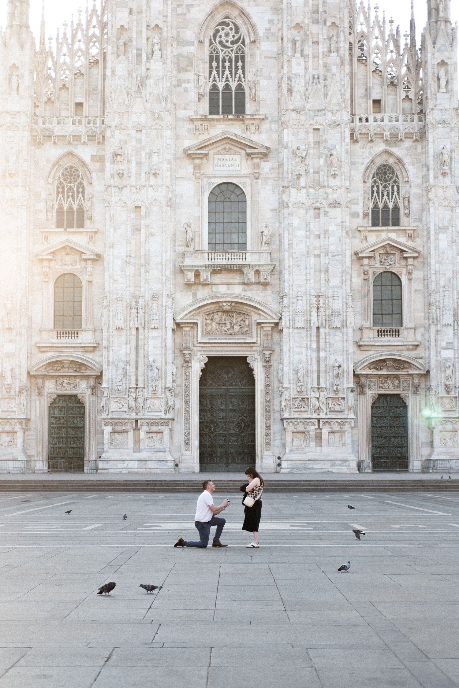 Man proposes to woman on one knee in Piazza del Duomo, Milan, with the Gothic cathedral facade behind them.
