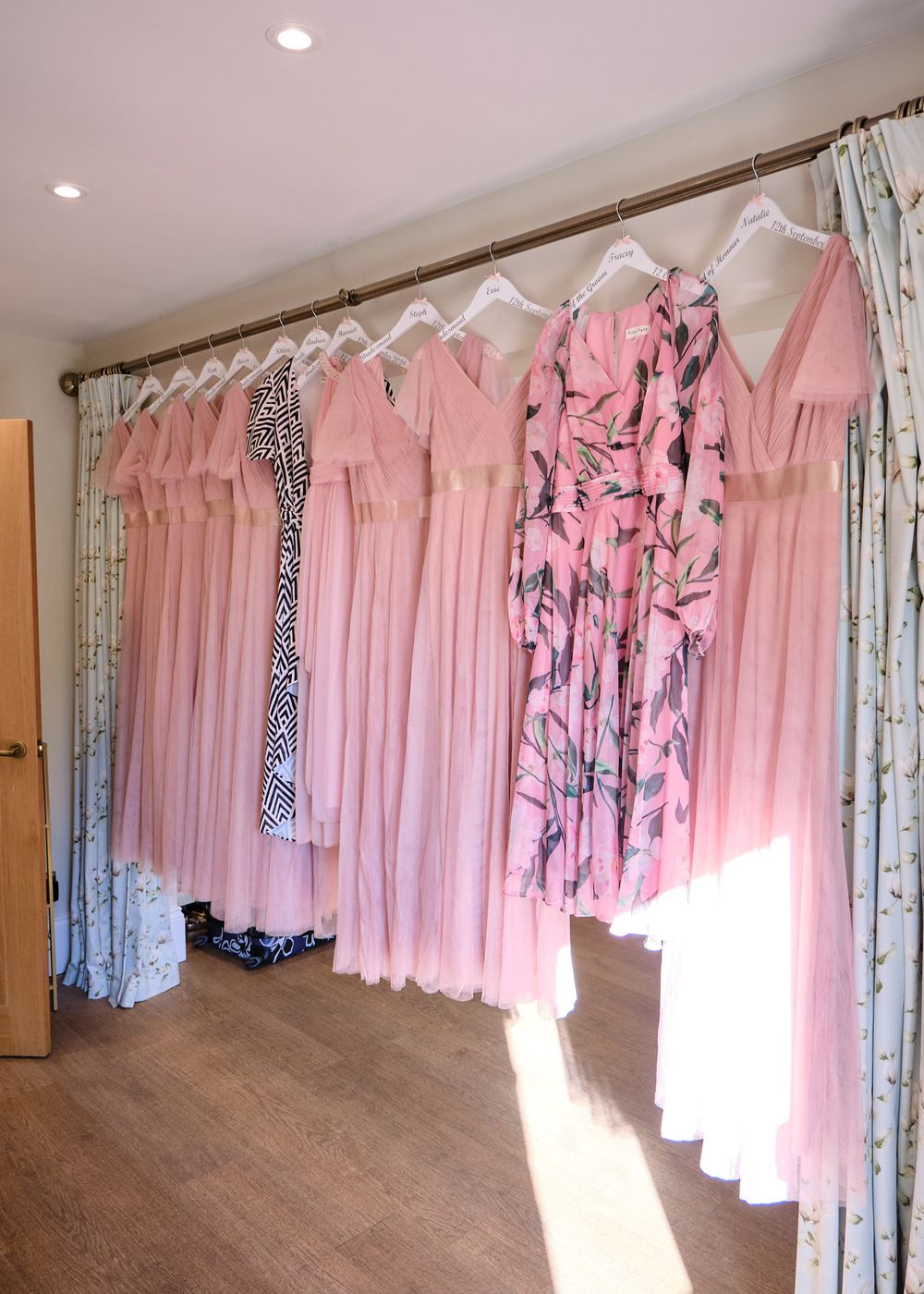 Pink bridesmaid dresses hanging on rail in sunlit dressing room before wedding.
