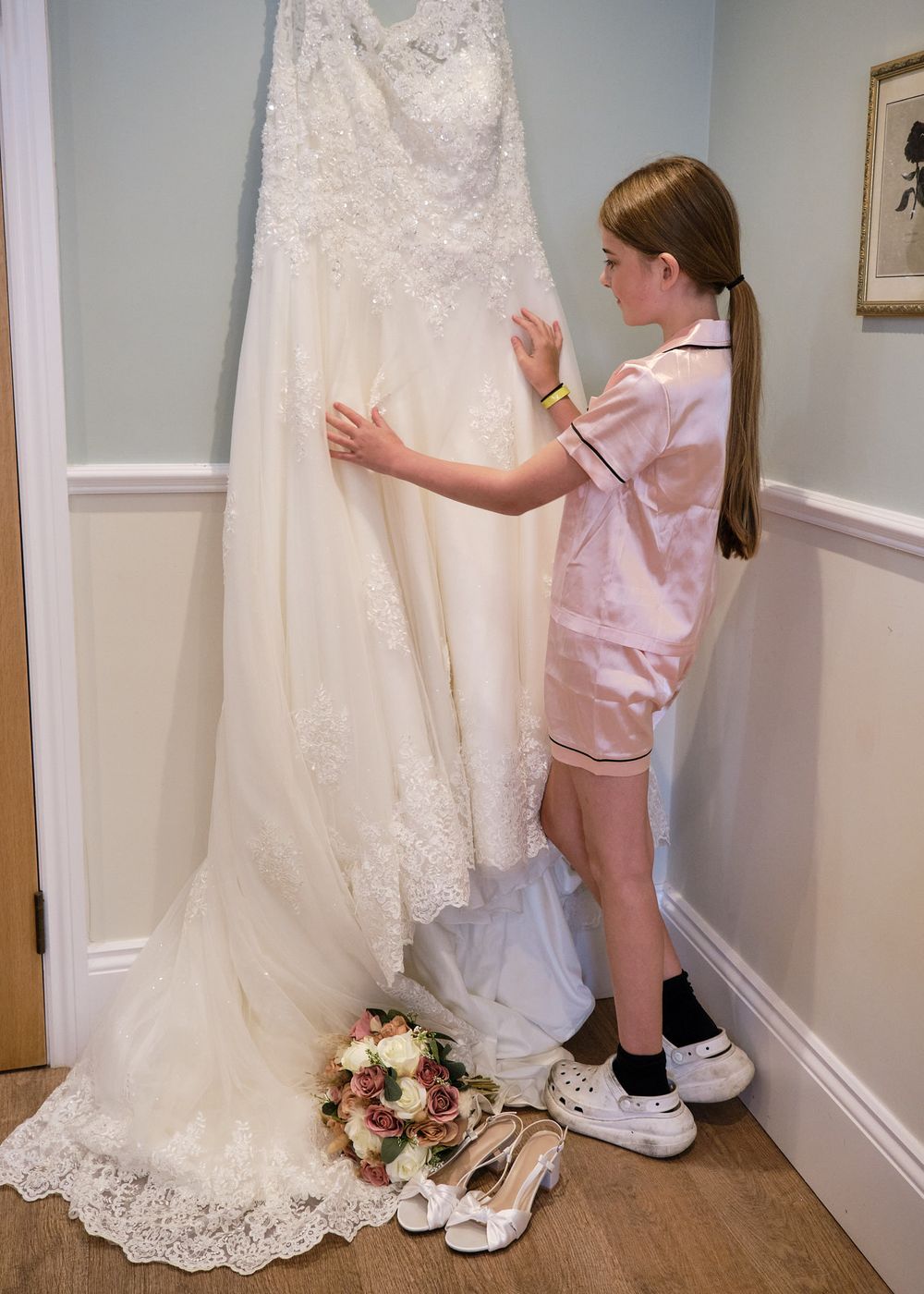 Young girl in pink pyjamas admiring lace wedding dress hanging with bridal bouquet.
