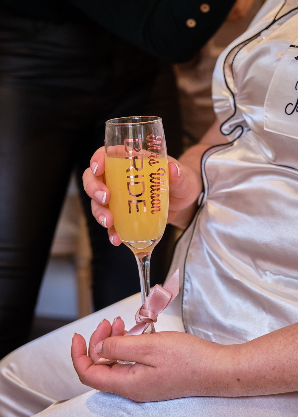 Close-up of a bride holding a champagne flute with gold text while wearing white satin wedding pajamas.