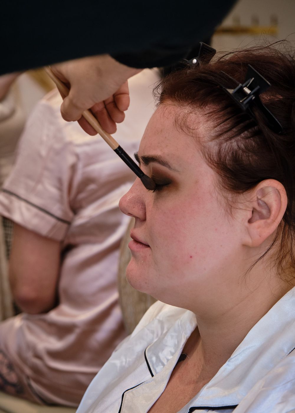 Professional makeup artist applying eye makeup with a brush to a young woman in pink satin pajamas.