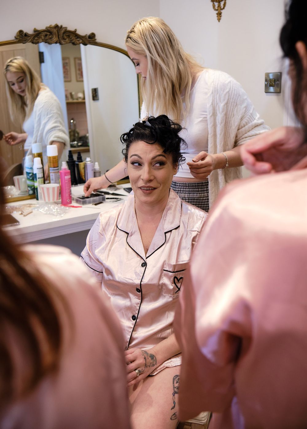 Bride in pink satin pajamas laughing with friends while getting hair styled in a bridal preparation room.