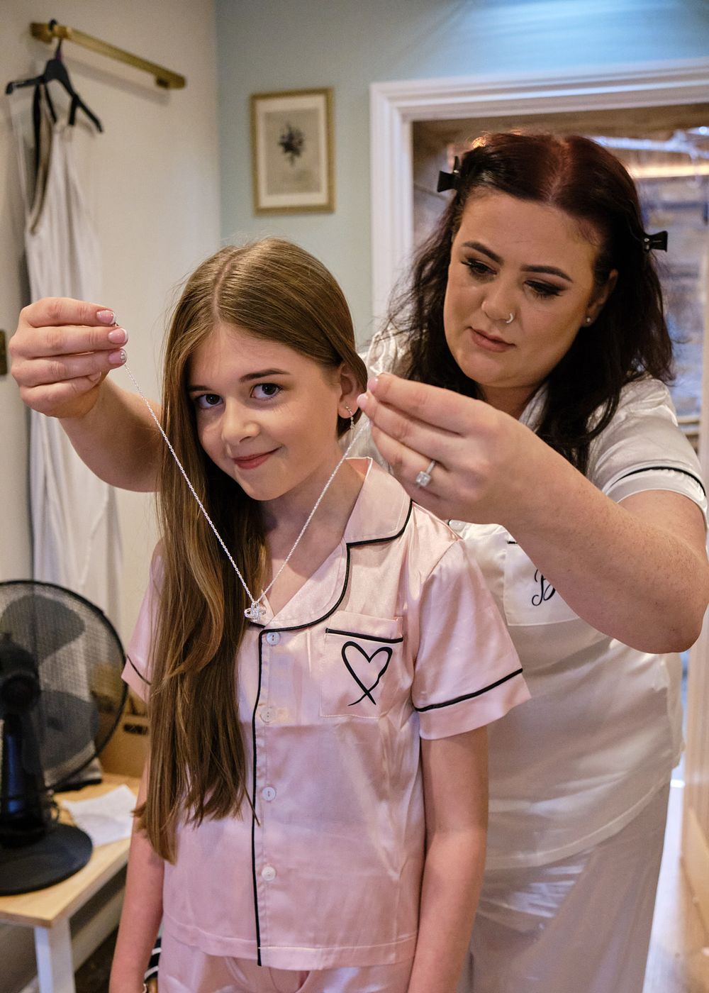 Woman in white satin pajamas helping a young girl put on a delicate necklace during wedding morning preparations.