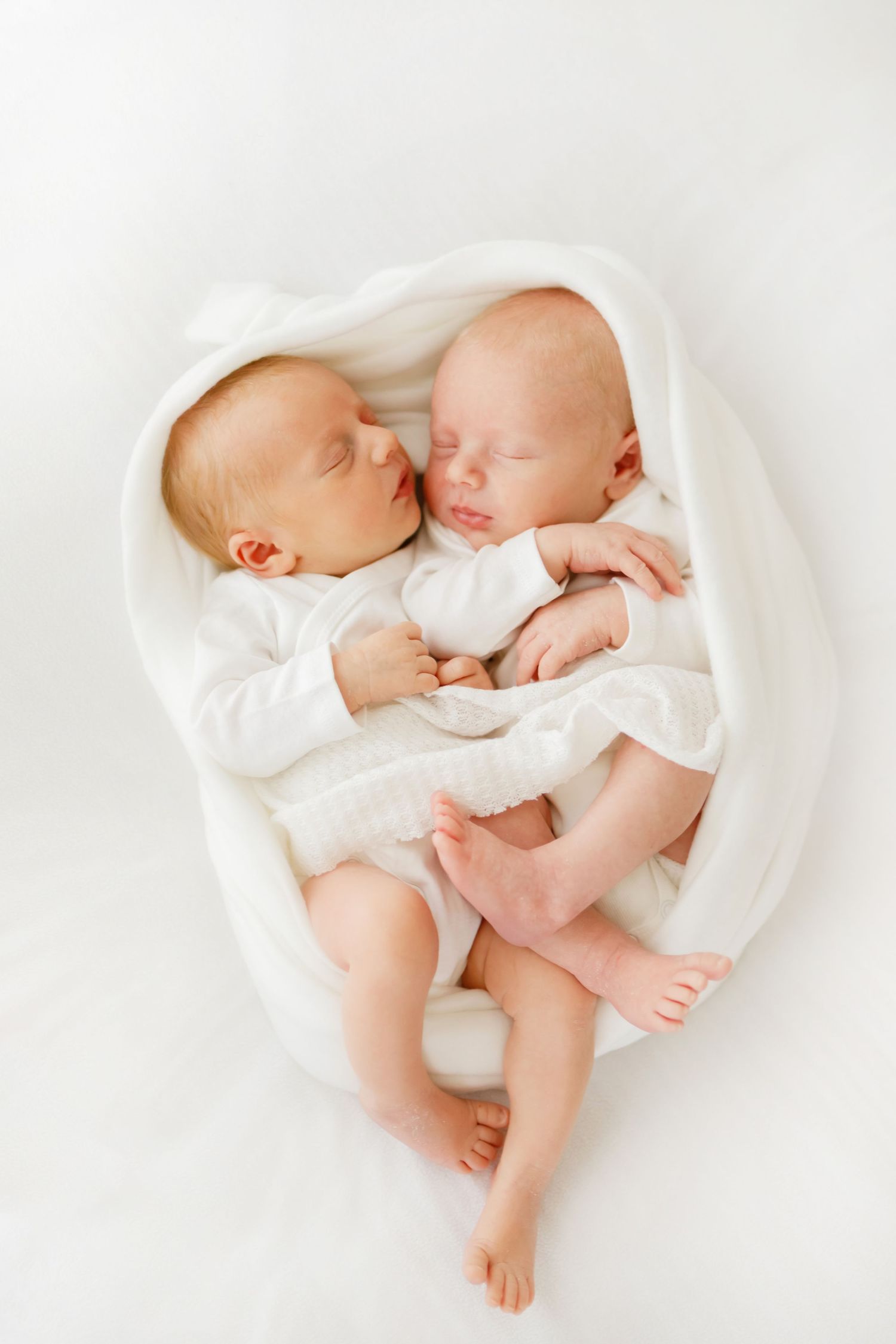 Two newborn twins sleeping peacefully together wrapped in a soft white blanket on a white background.