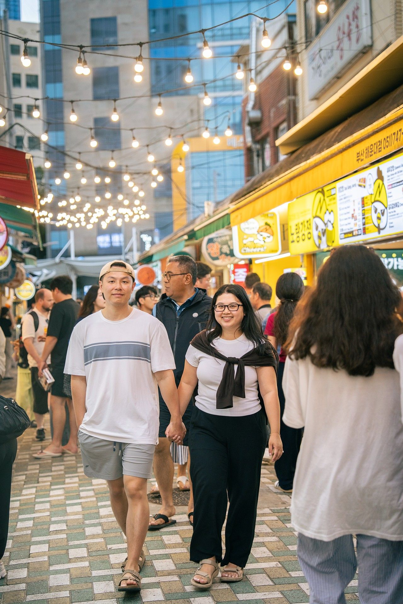 Couple holding hands walking through a busy Korean street market at night with string lights and food stalls.