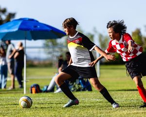 Two youth soccer players compete for the ball during an outdoor match, one in white and one in red checkered jersey.
