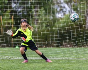 Female soccer goalkeeper in neon yellow jersey diving to block a shot during a match on a green field.