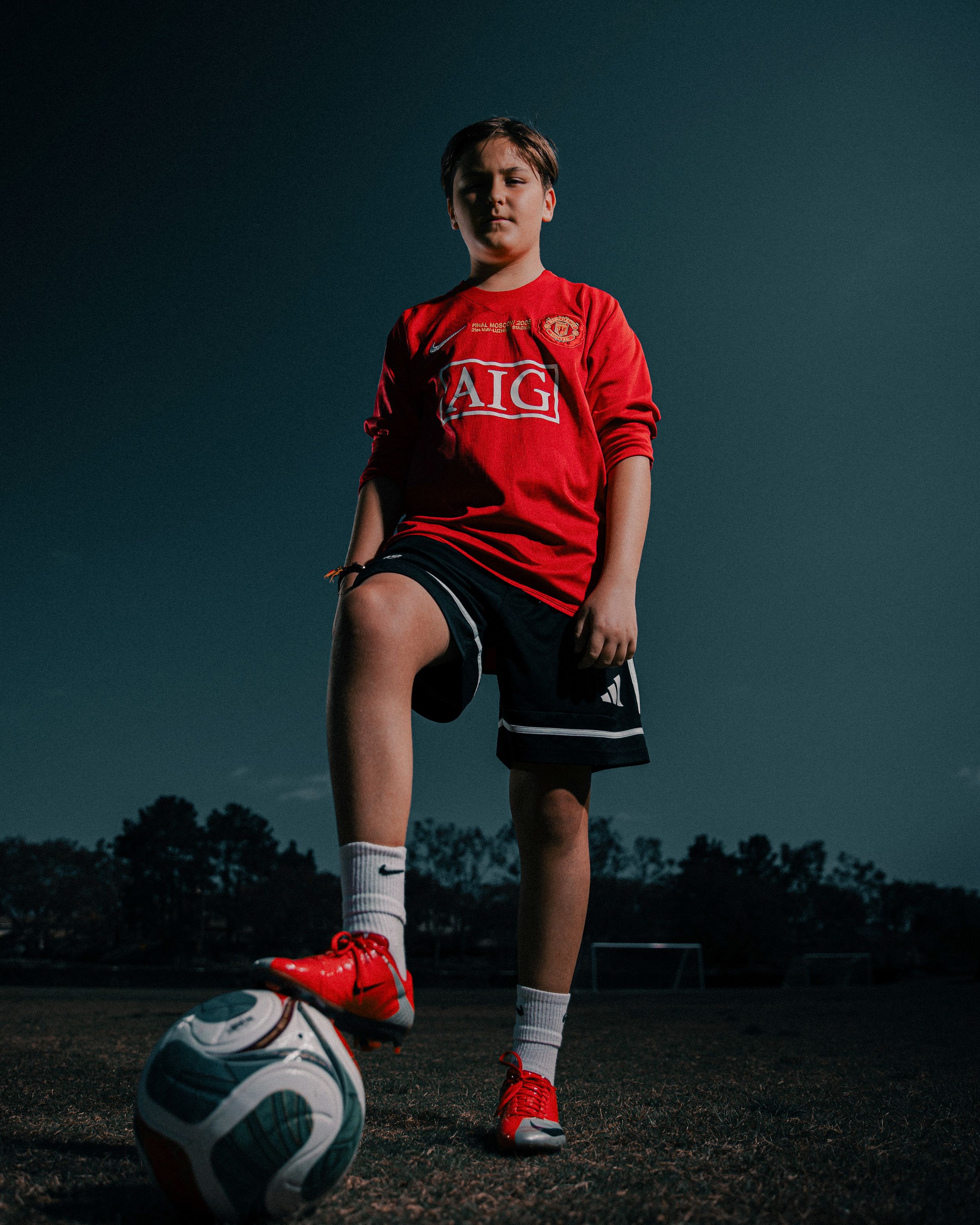 Young soccer player in red Manchester United AIG jersey poses with foot on ball outdoors at dusk.