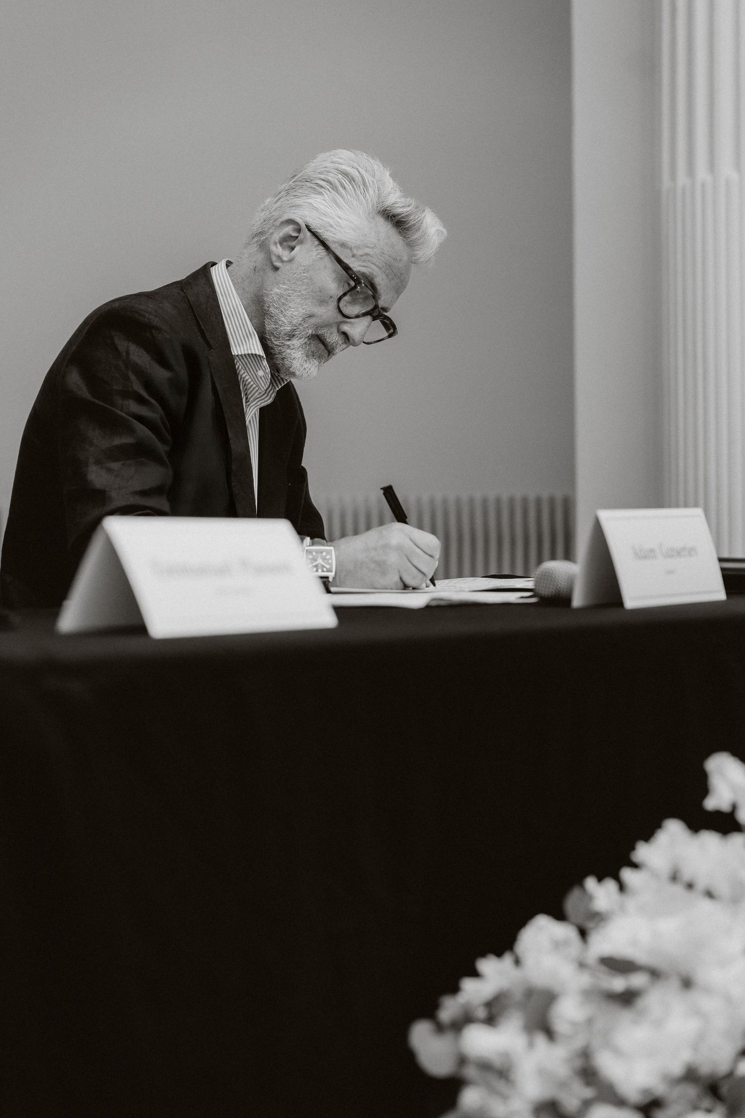 Elderly man with glasses signing documents at a formal table in black and white photography.