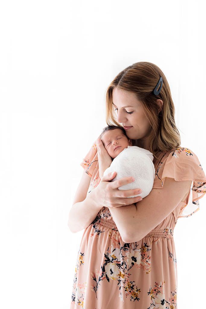A mother in a floral dress tenderly holds her swaddled newborn baby close to her chest against a white background.