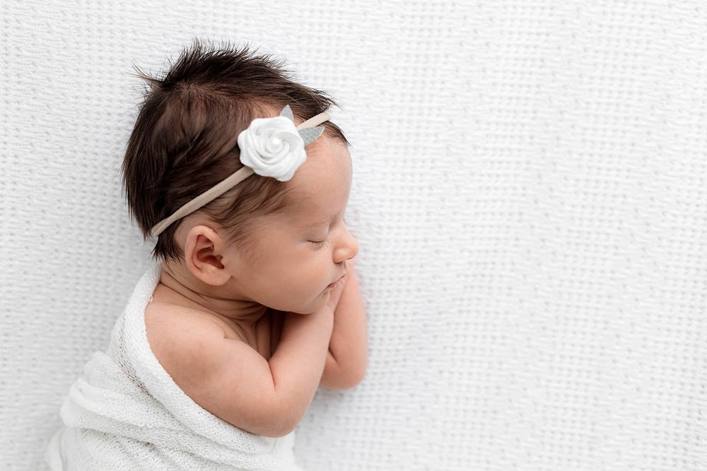 Newborn baby girl sleeping peacefully wrapped in white blanket wearing a delicate white flower headband.