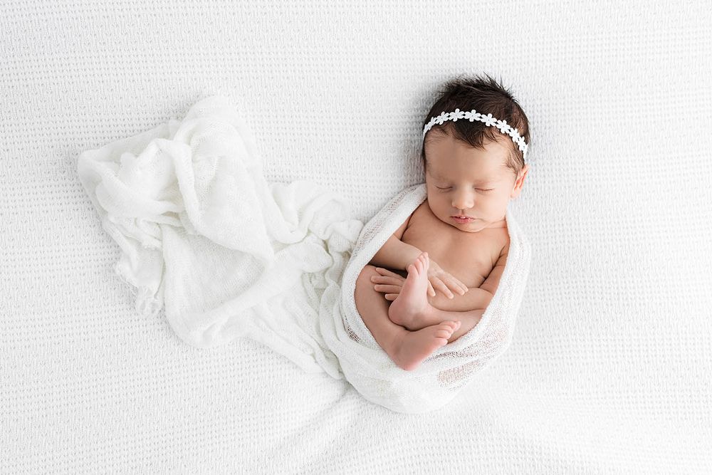 Sleeping newborn baby girl wrapped in white swaddle with floral headband on white background by Menifee newborn preemie photographer.
