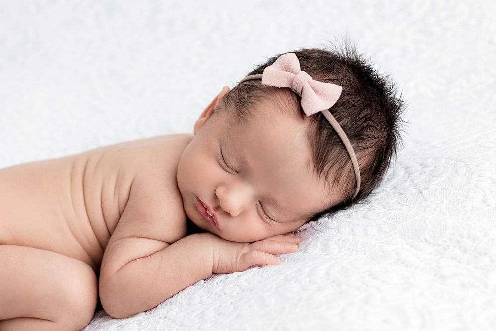 Sleeping newborn baby girl with dark hair wearing a pink bow headband on a white fluffy blanket.
