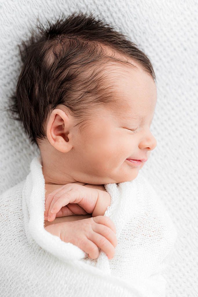 Peaceful sleeping newborn baby with dark hair wrapped in a soft white knit blanket, smiling gently.