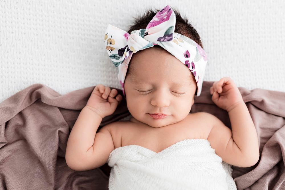 Sleeping newborn baby girl wearing a floral bow headband, wrapped in white, lying on a soft mauve blanket.