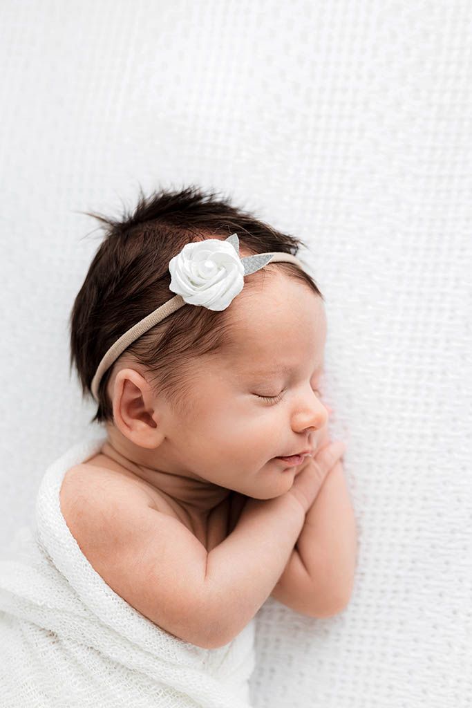 Sleeping newborn baby girl with dark hair wearing a white rose headband by Menifee newborn preemie photographer.