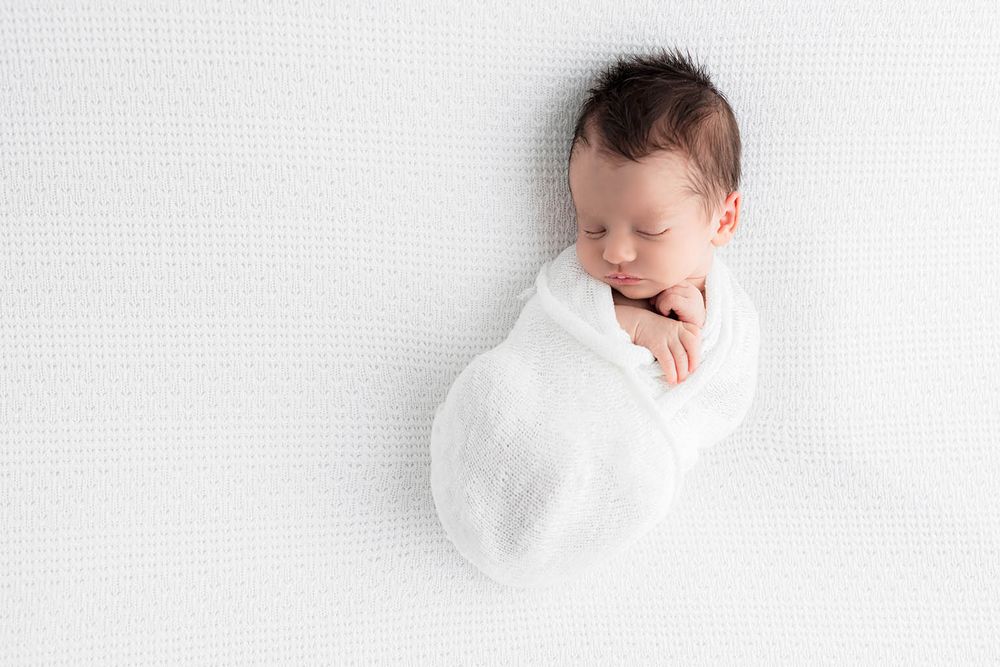 Peaceful newborn baby swaddled in white wrap sleeping on white textured blanket, professional newborn photography.