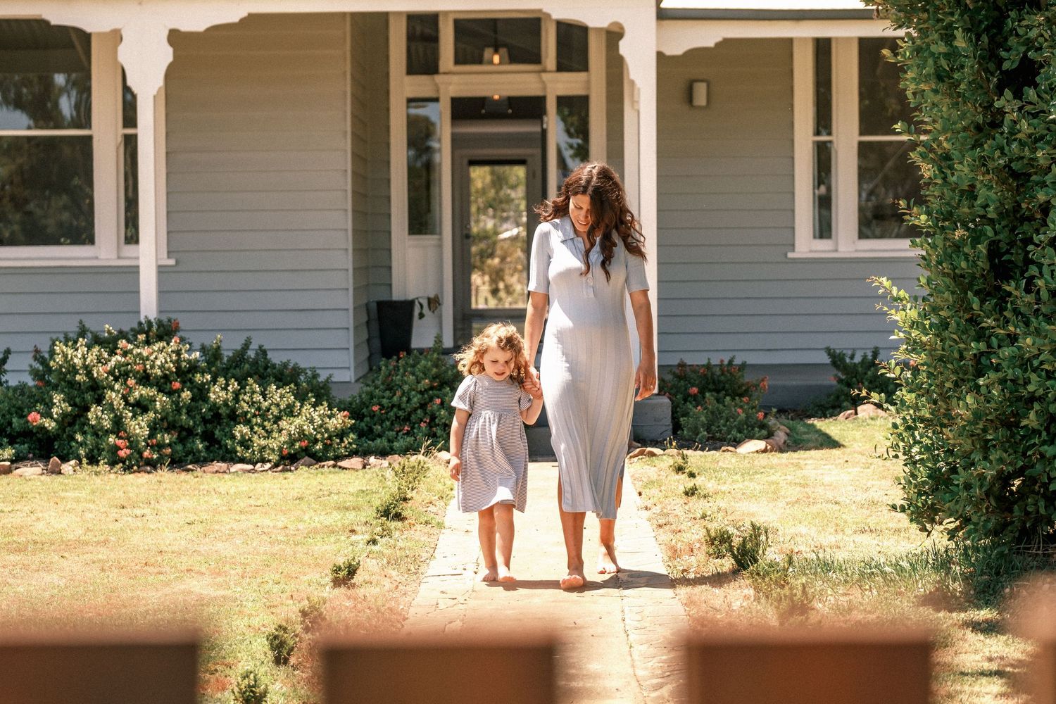 A mother and young child walk hand-in-hand along a brick path in front of a charming grey house.