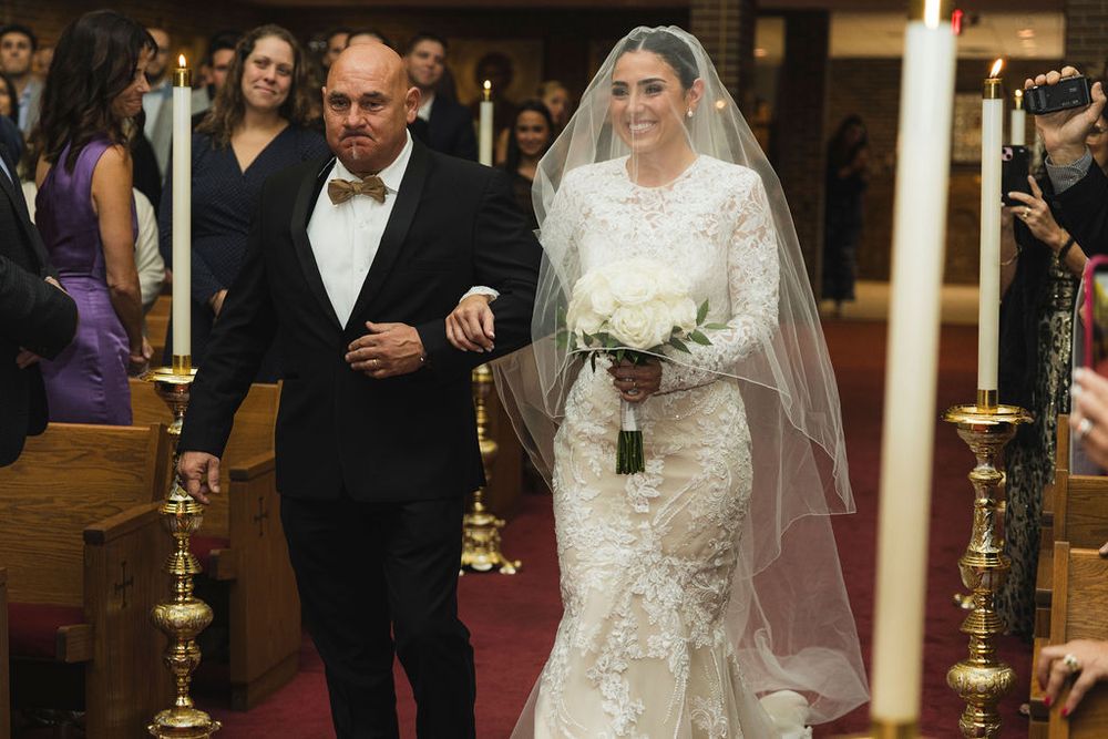 Bride in lace gown and veil walks down church aisle with her father, holding white bouquet at elegant wedding ceremony in Chicago