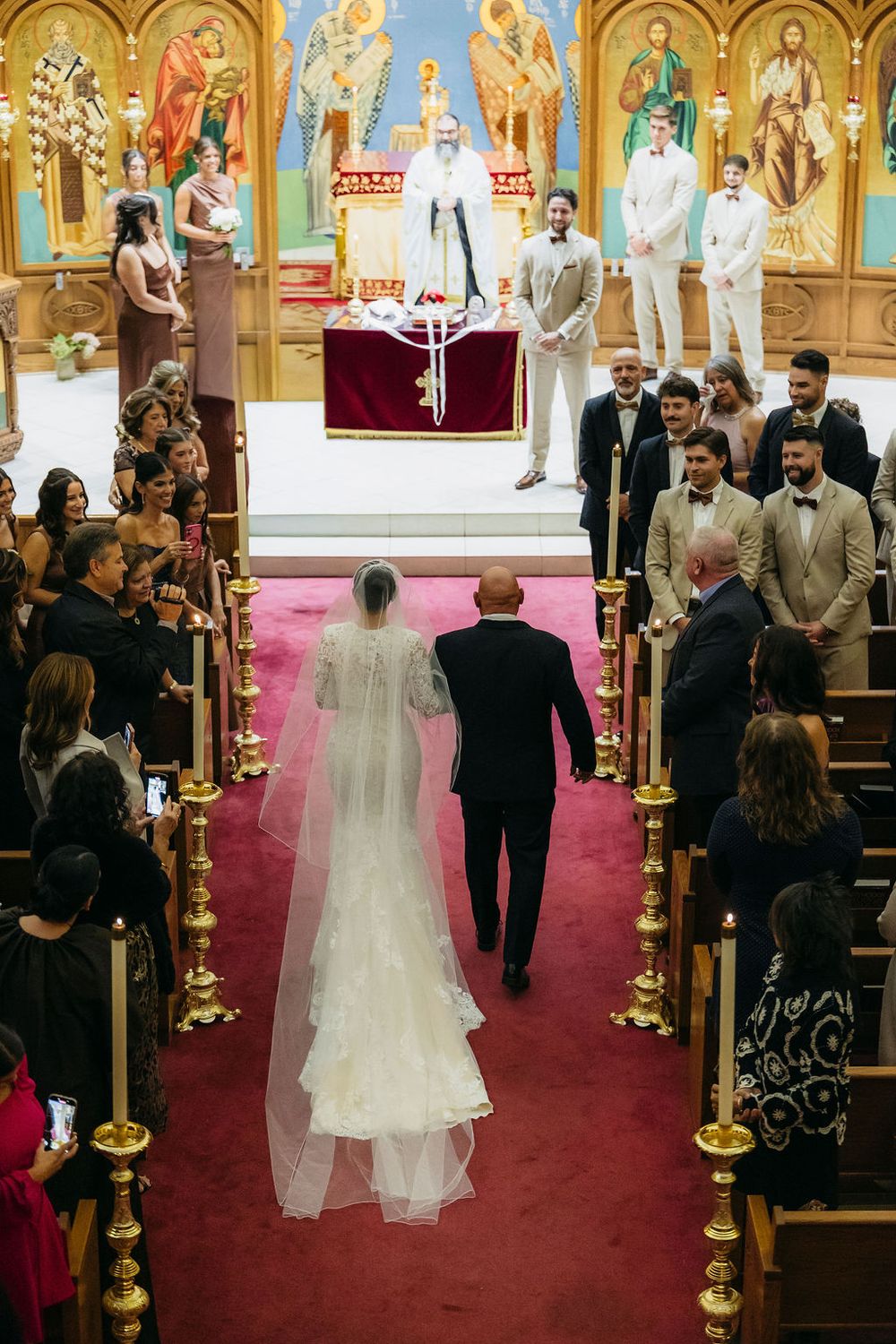 Bride walks down red-carpeted Chicago church aisle with her father toward altar, guests and groomsmen watching in ornate Orthodox church.