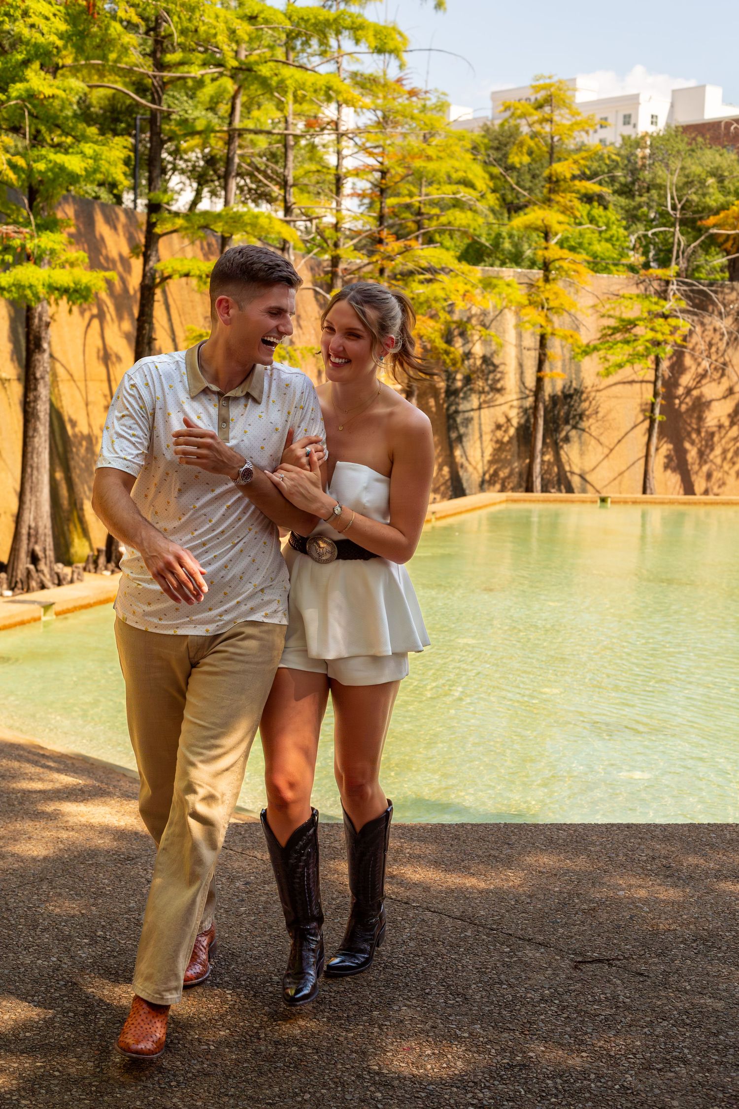 Happy couple walking together near a scenic pool, woman in white dress and cowboy boots, man in casual attire.