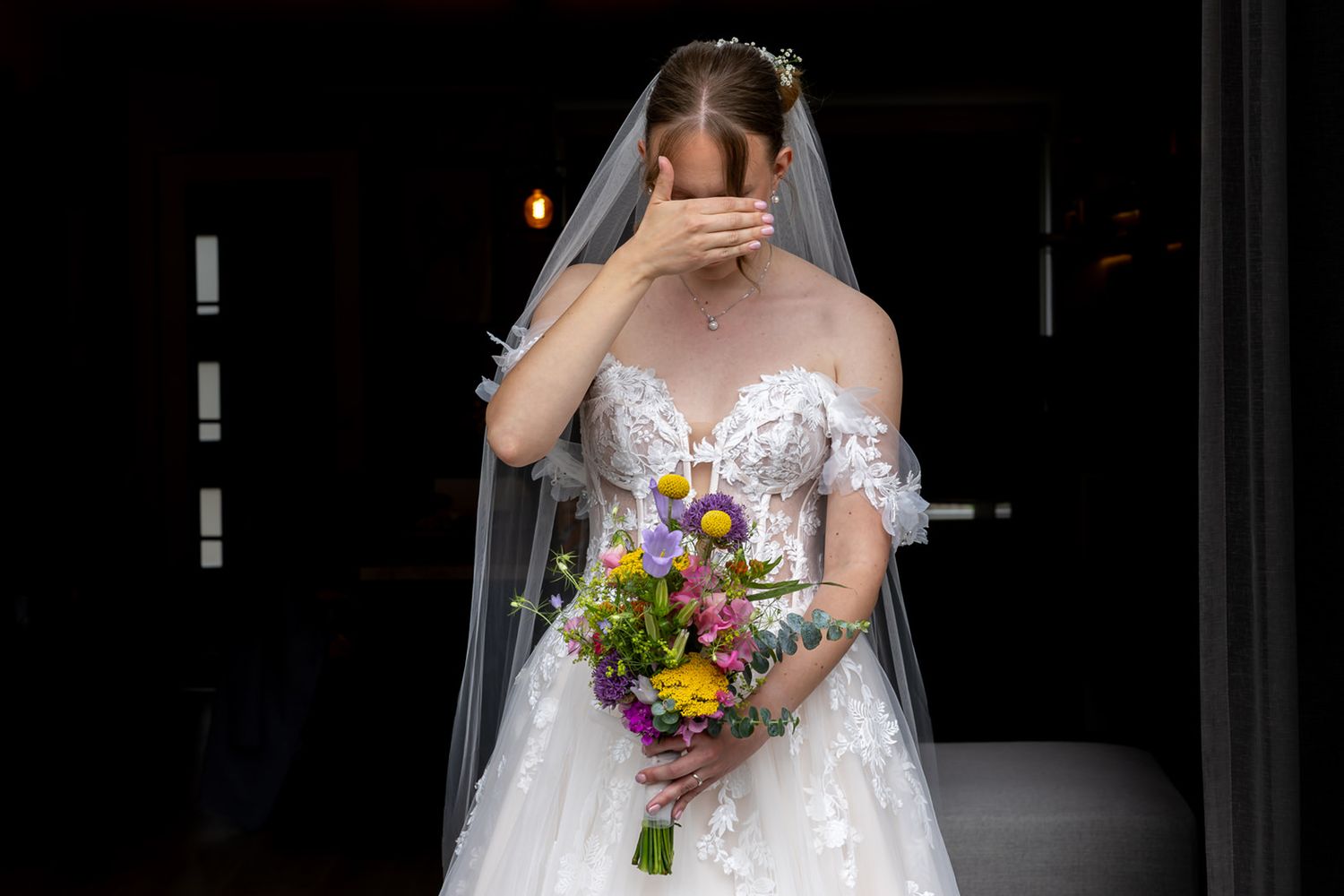 Bruid staat in de deuropening van de tuindeuren in haar trouwjurk met kleurrijk bruidsboeket, handen voor de ogen tijdens het first look