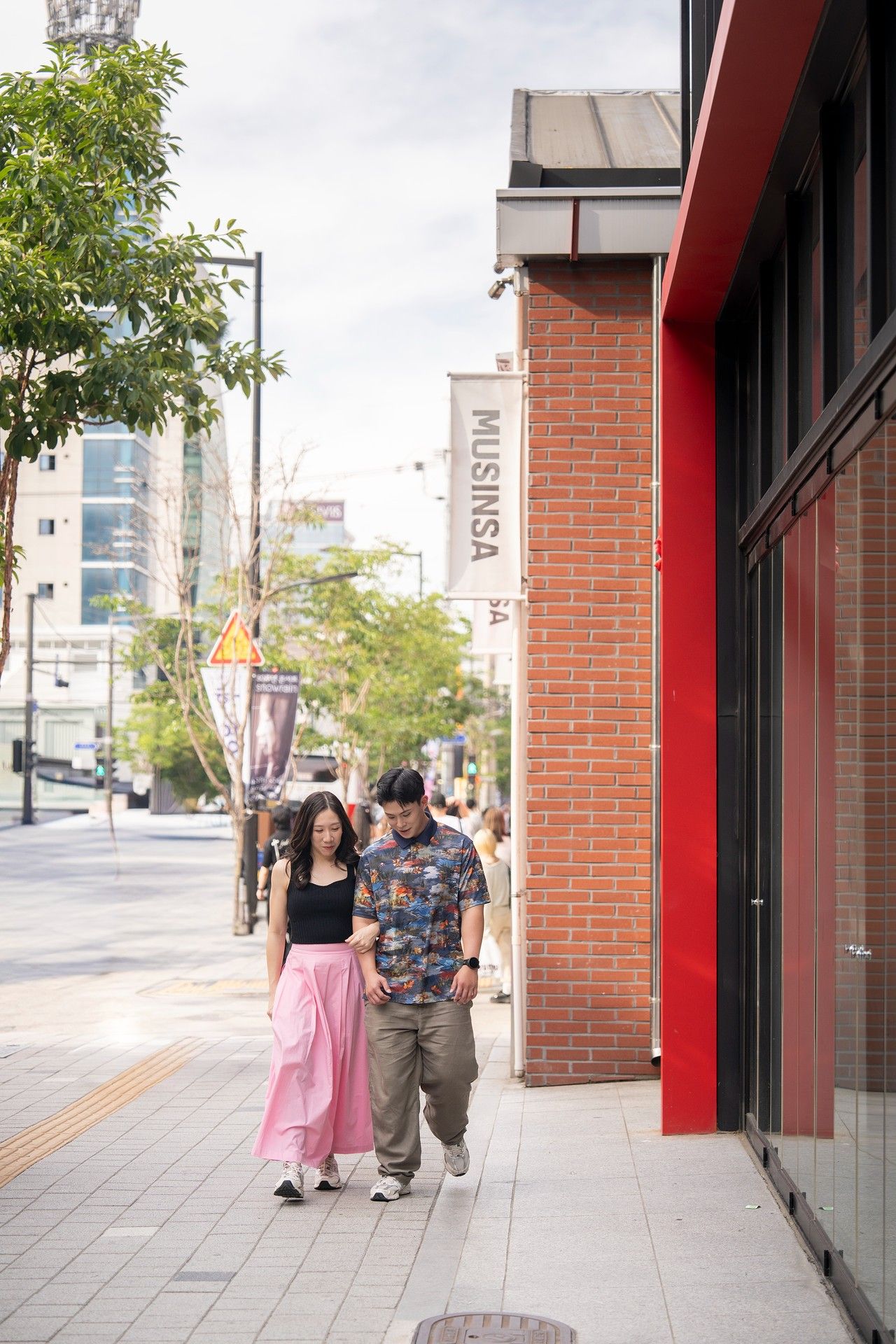 Couple walking together on a sidewalk near a Musinsa store in an urban city street setting.