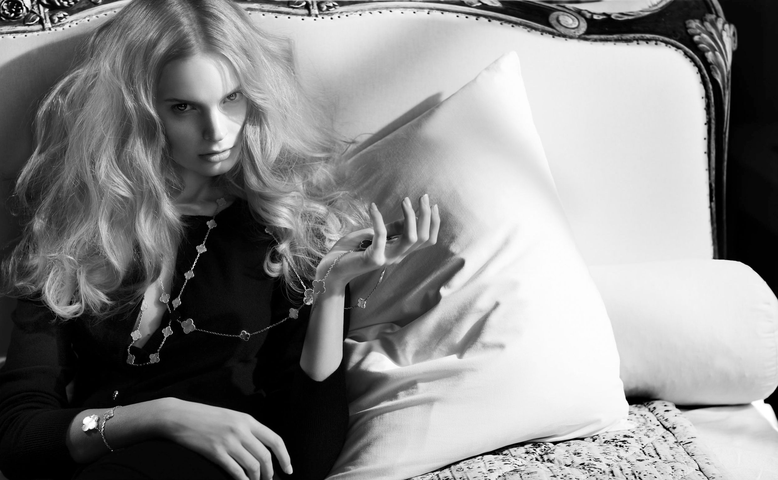 Black and white photo of a blonde woman reclining on an ornate sofa, wearing a dark top with a long necklace.