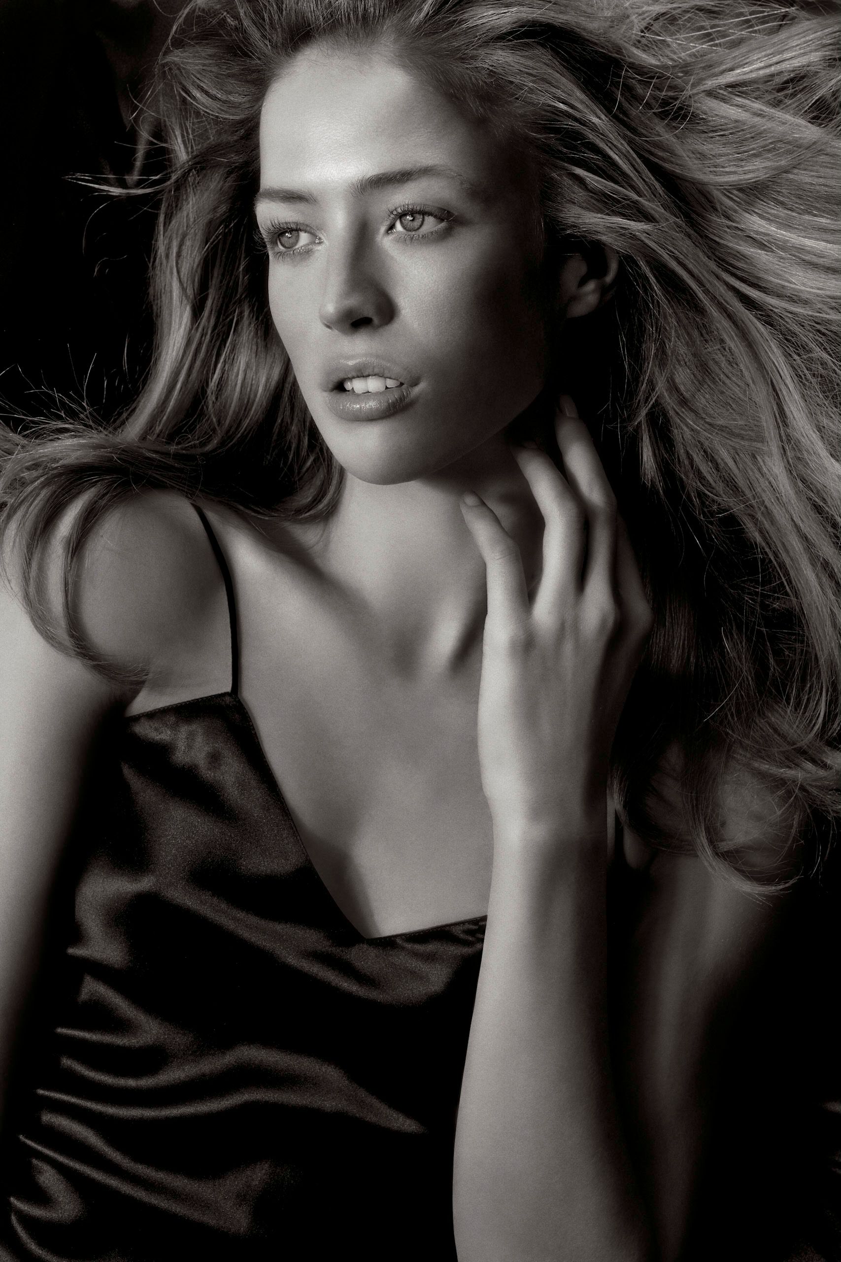 Black and white portrait of a young woman with flowing hair wearing a dark satin dress, hand raised to her neck.
