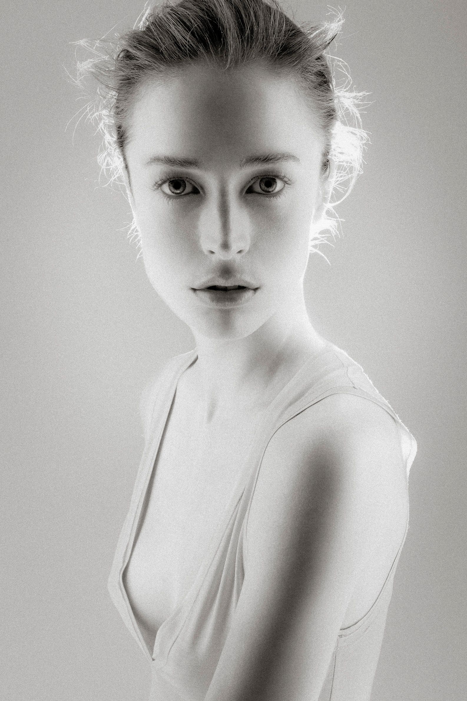 Black and white studio portrait of a young woman in a white dress with hair up, gazing directly at camera.