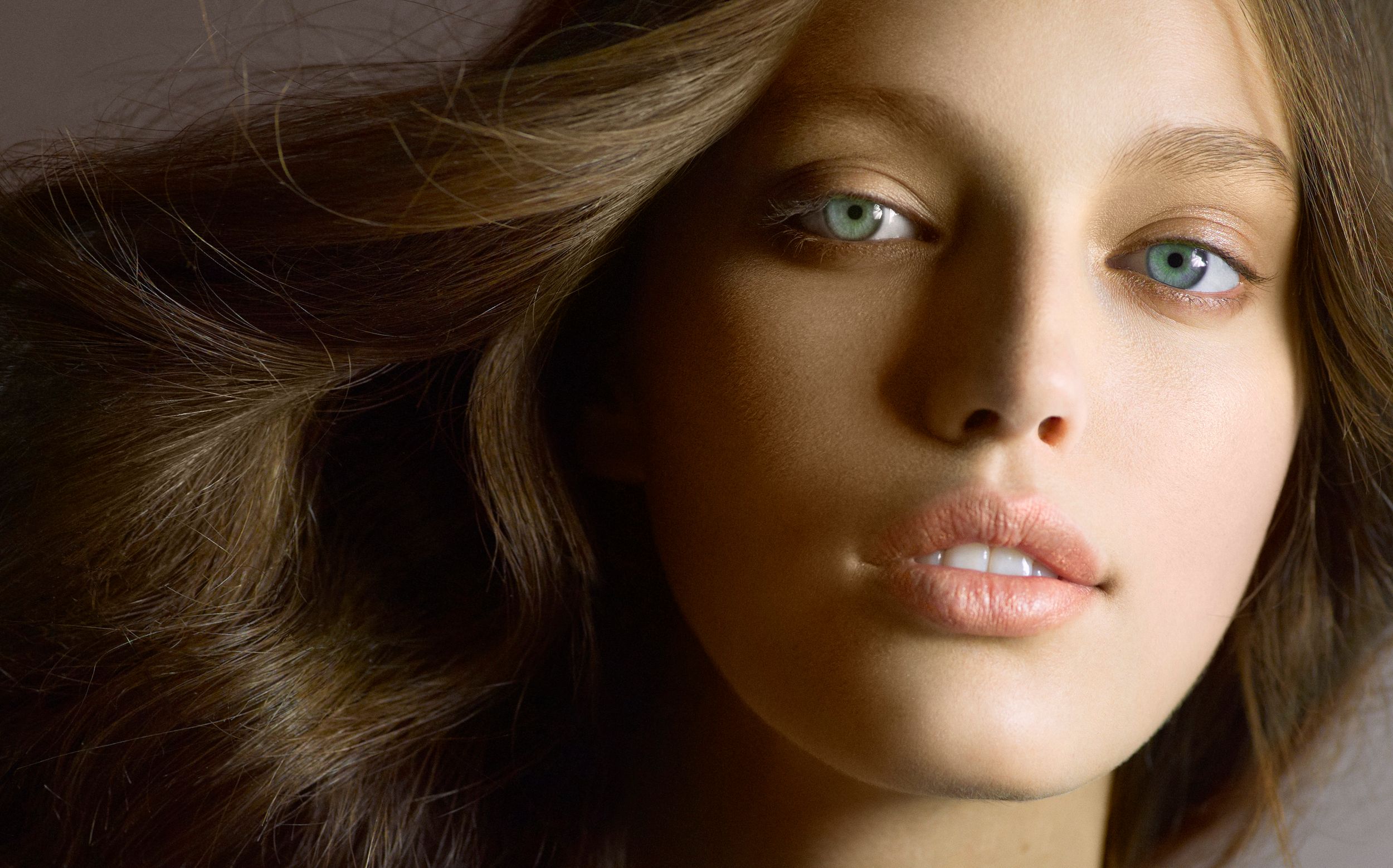 Close-up portrait of a young woman with blue eyes and flowing brown hair against a neutral background.