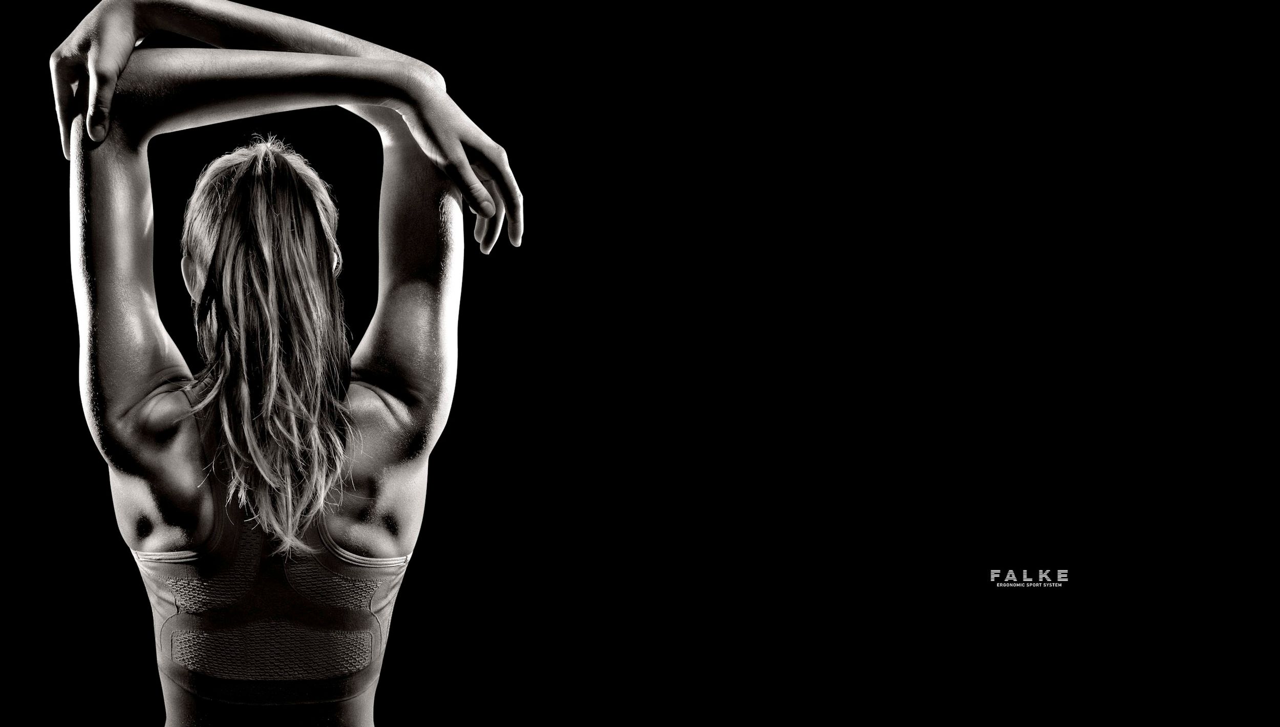 Black and white photo of a fit woman stretching her arms overhead, showcasing toned back muscles against dark background.