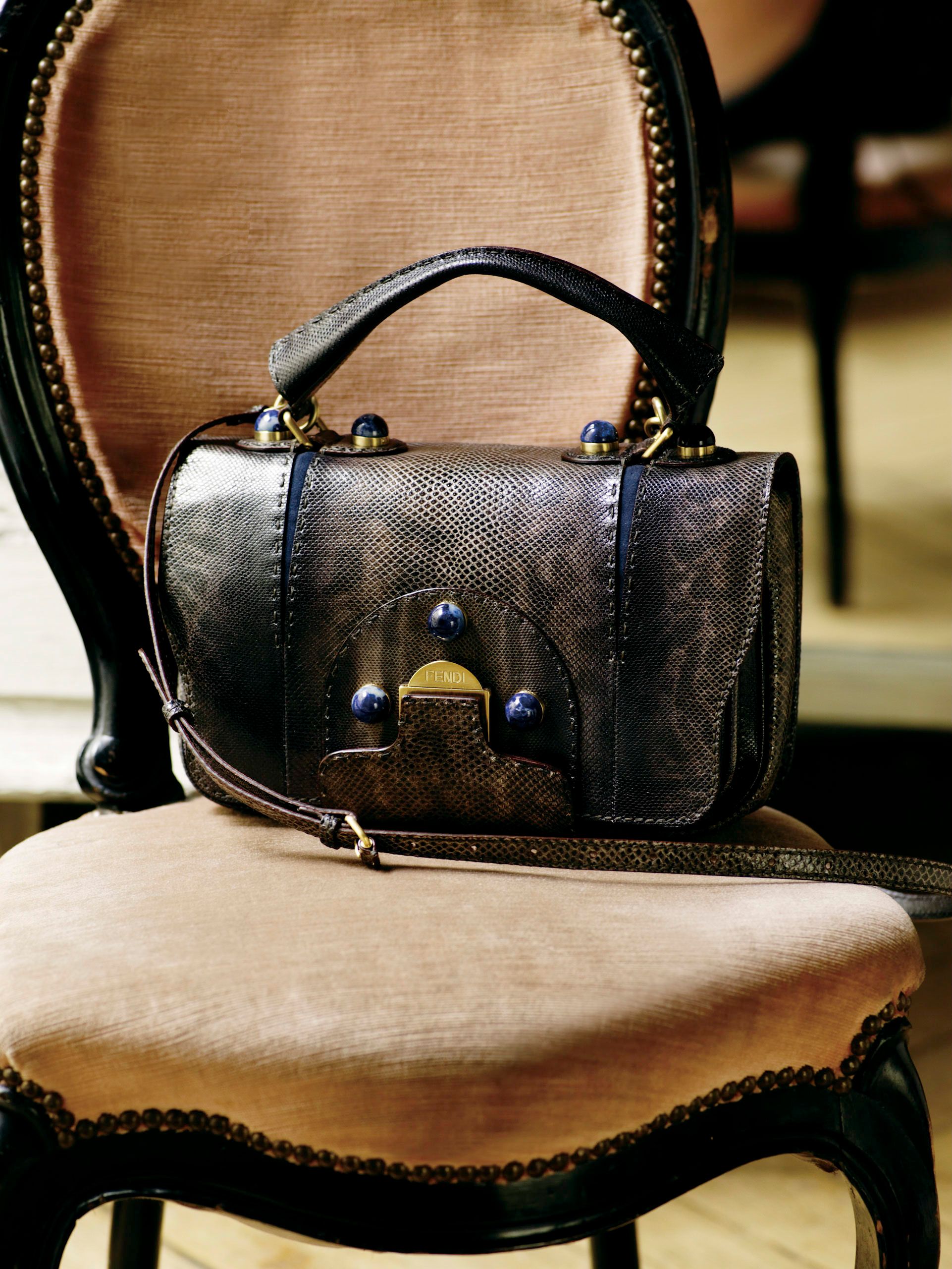 Dark exotic leather structured handbag with gold hardware and blue accents resting on a vintage velvet chair.