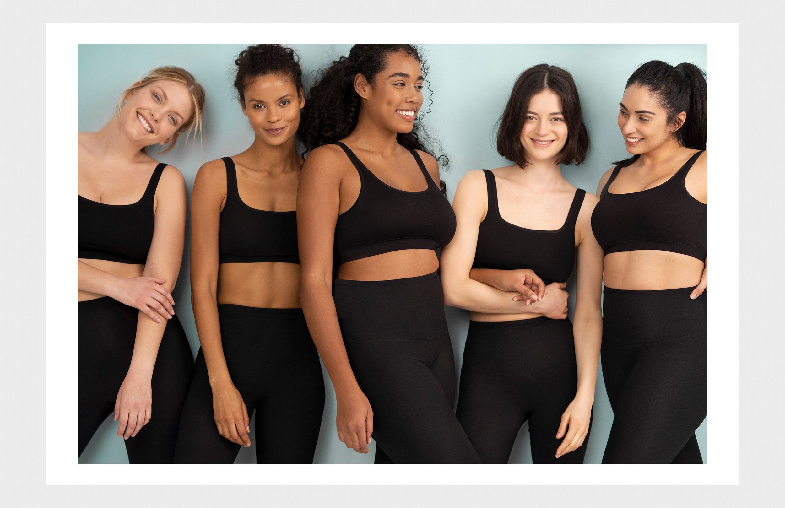 Five diverse women in matching black sports bras and leggings smiling together against a light blue background.