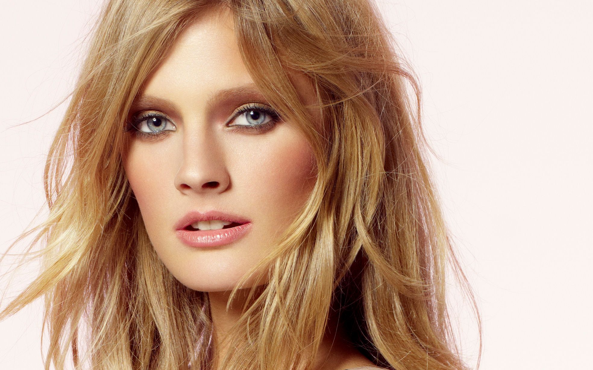 Close-up portrait of a young woman with tousled blonde hair and blue eyes against a soft white background.