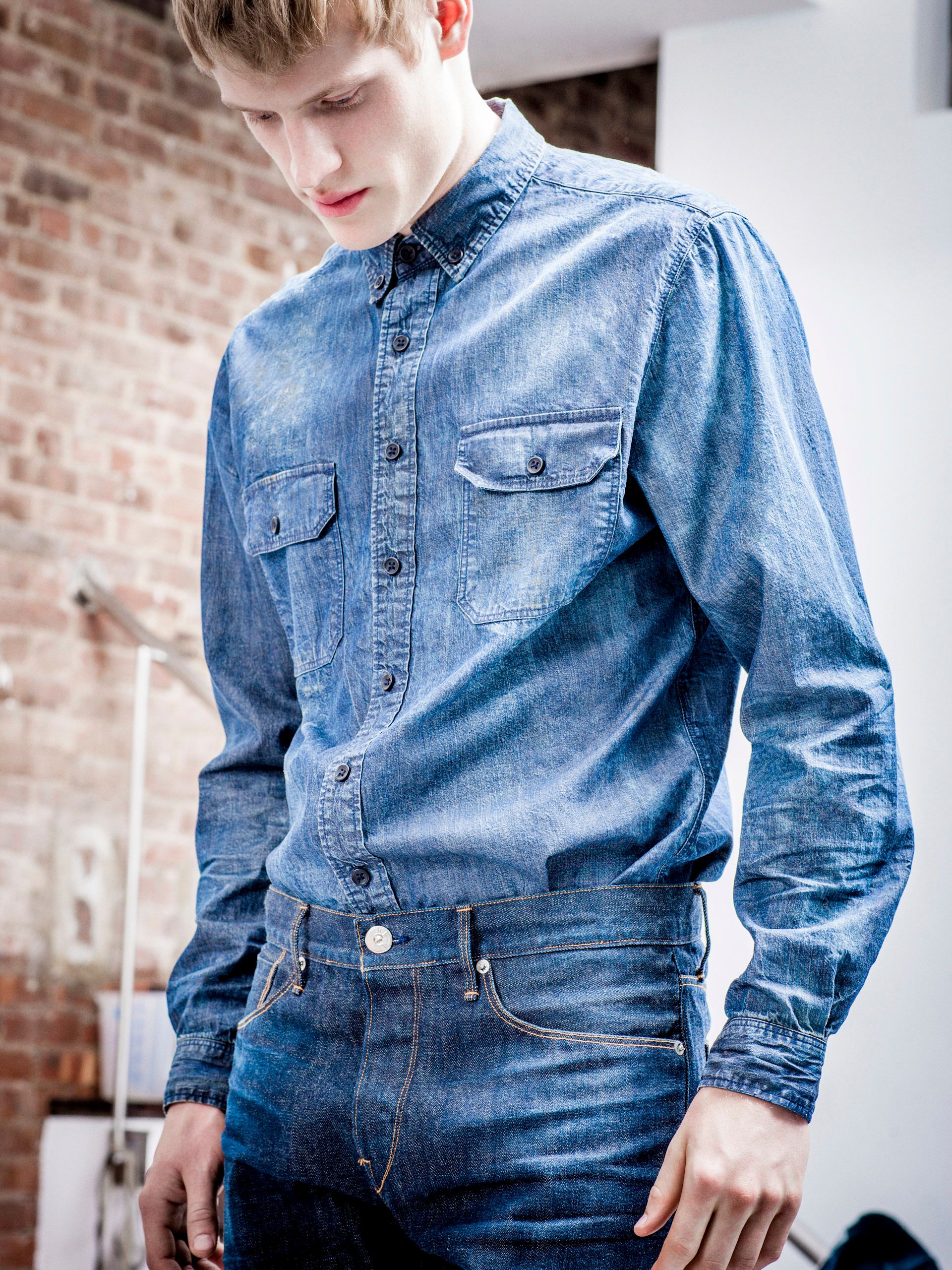 Man wearing a double-pocket blue denim chambray shirt paired with dark wash jeans in a loft setting.