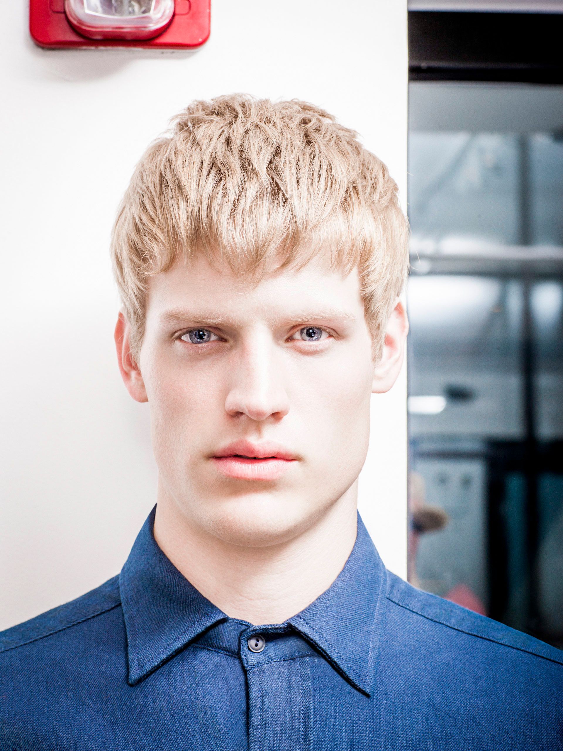 Young blonde man with blue eyes wearing a denim shirt, posing indoors near a white wall.